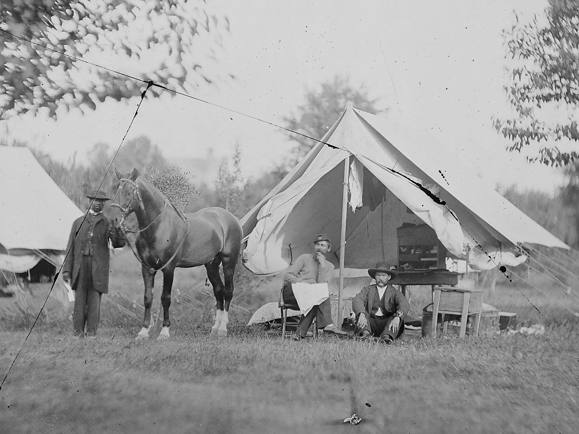 Two men sitting in a tent and a man standing holding a horse